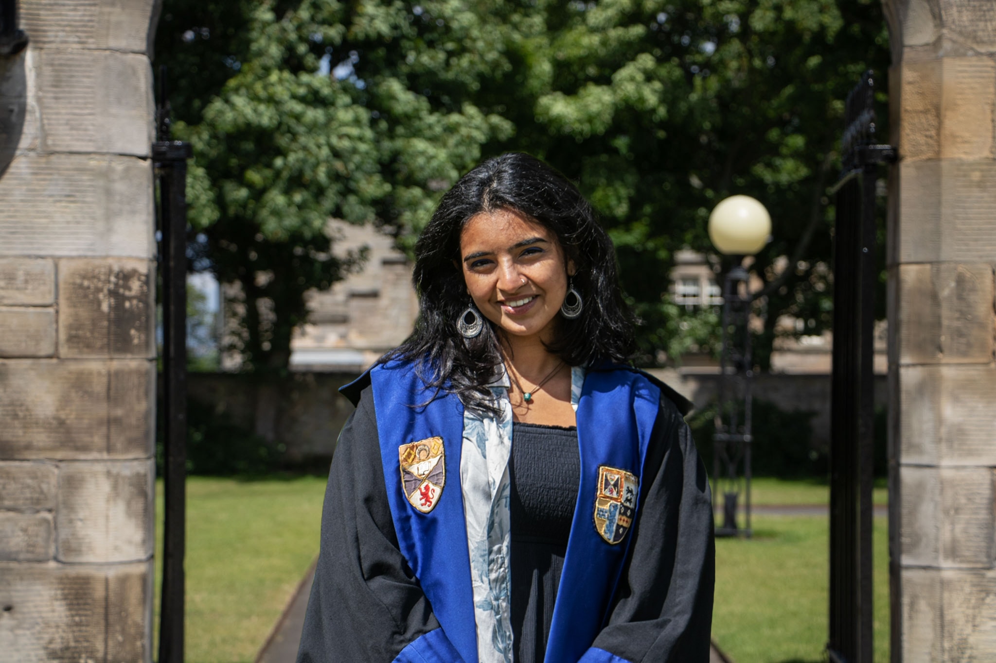 Hitanshi smiles in her black gown. The gown is faced in blue with the Association and University crests emblazoned.