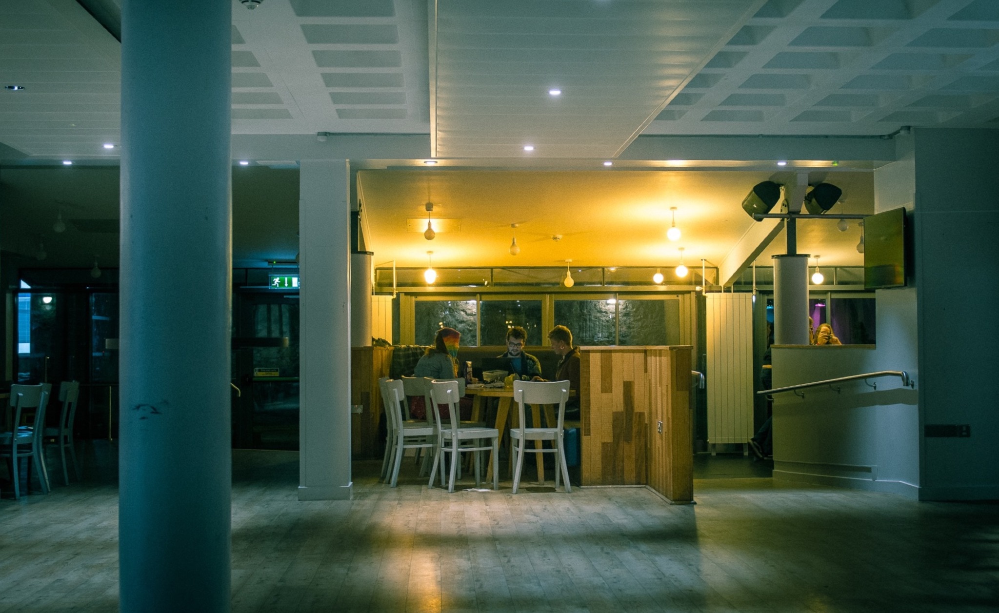 Three students sit working in Main Bar. It's dark, they are backlit by lights on the ceiling.