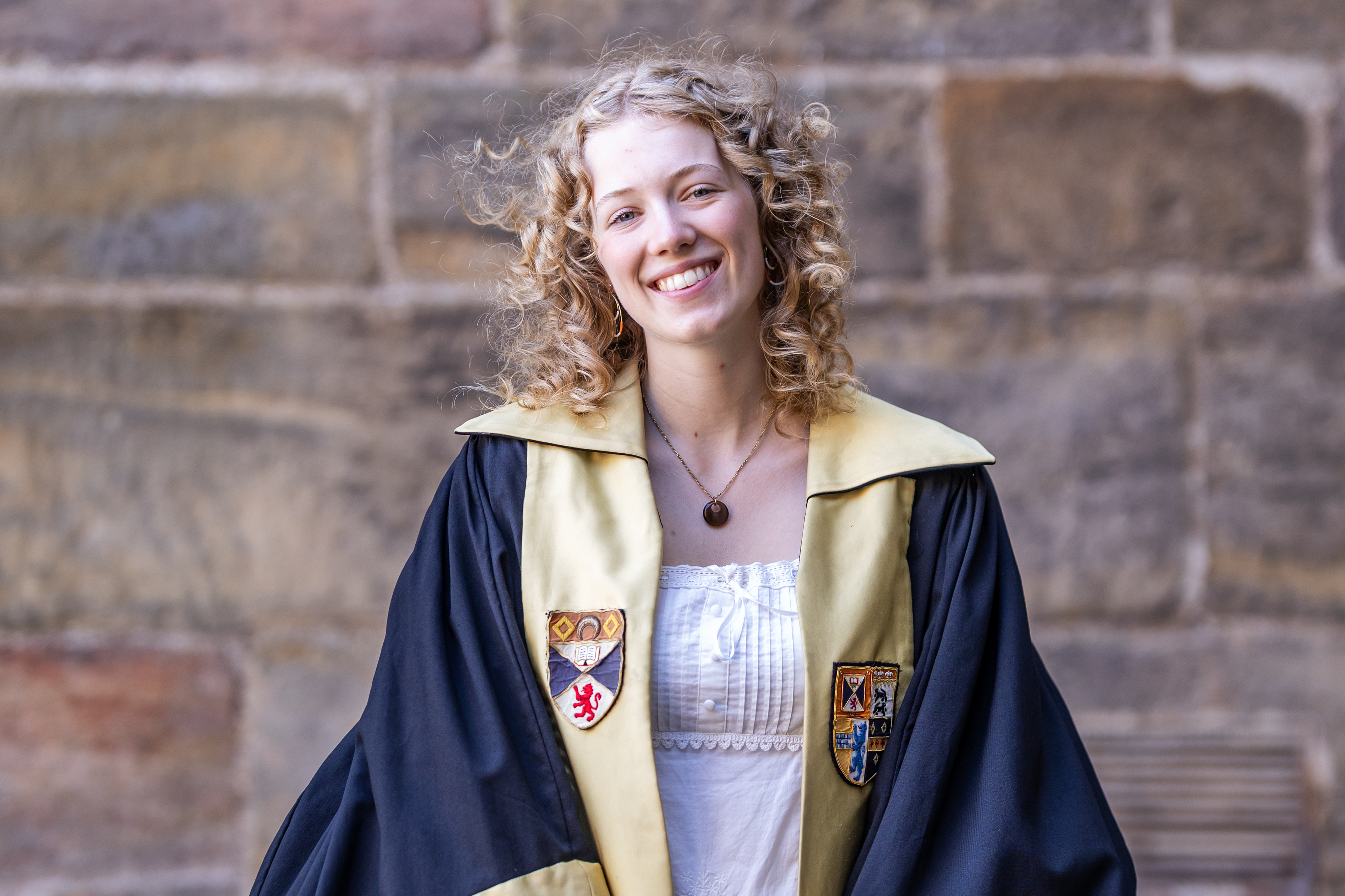 Emily smiles in the President of Education gown. The gown is faced in blue with the Association and University crests emblazoned.