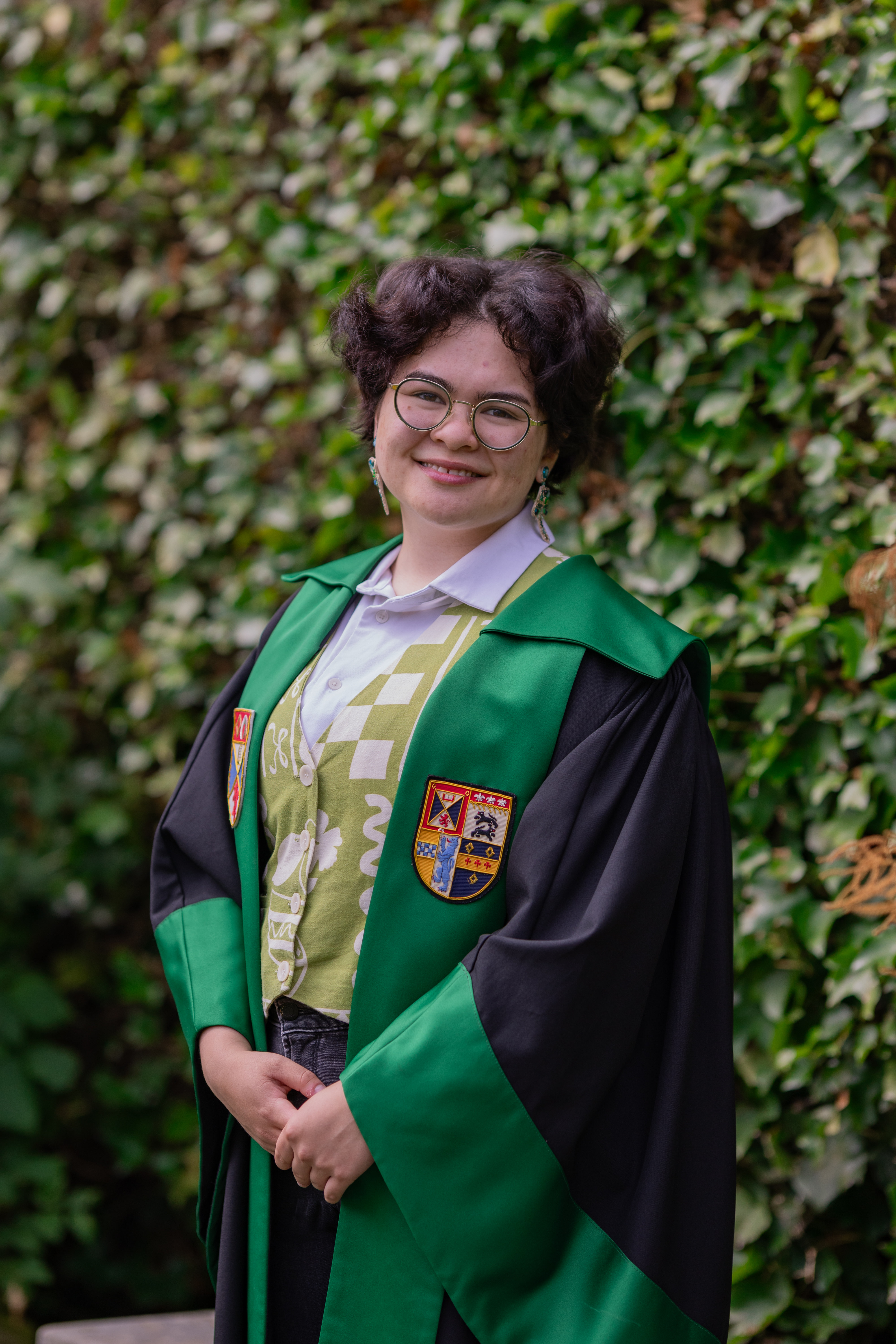 Alex smiles in the President of Wellbeing &amp; Community gown. The gown is faced green with the Association and University crests emblazoned.