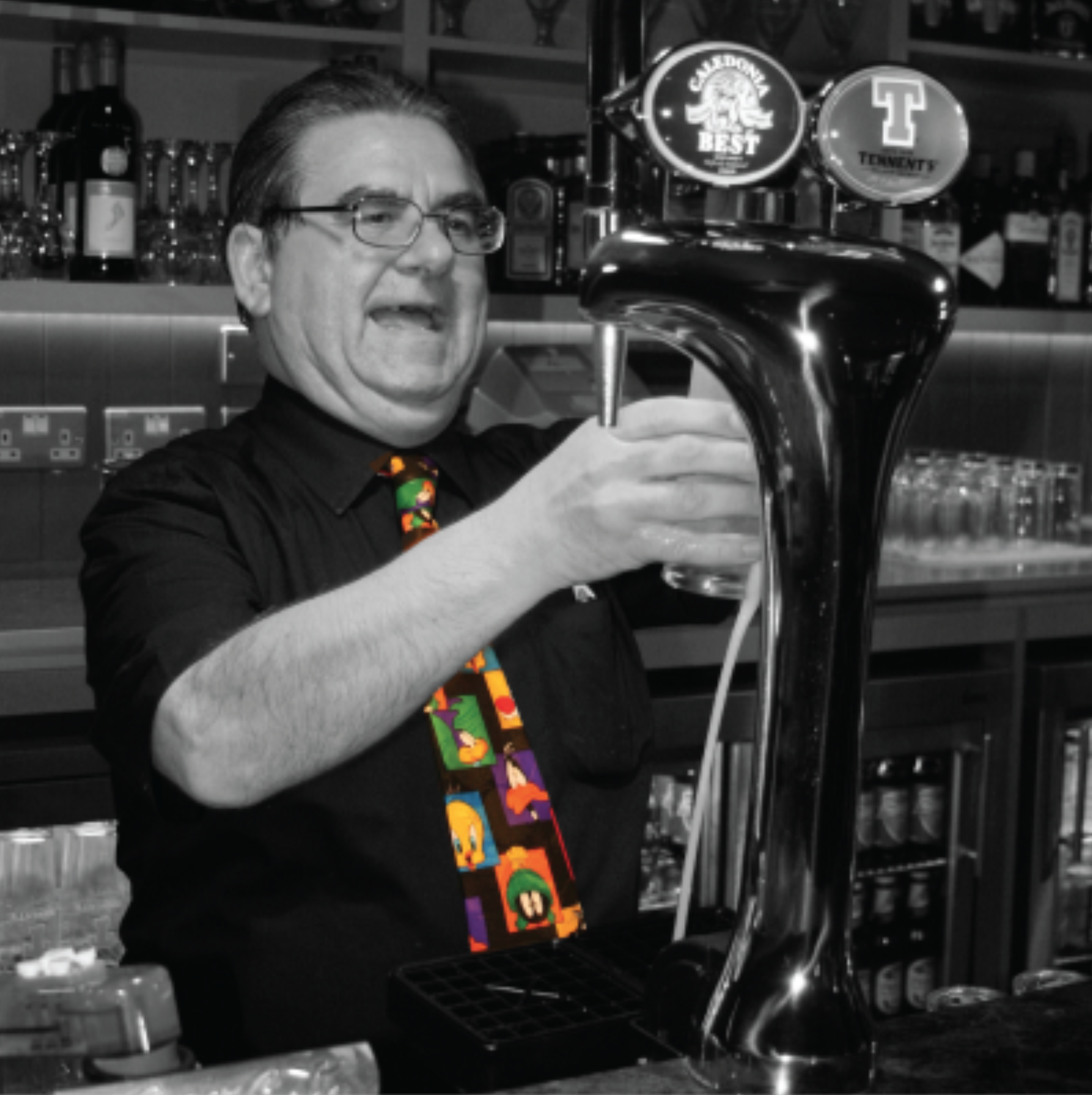 Sandy pulls a pint at the bar, he is smiling and wearing a tie with images of Looney Tunes characters.