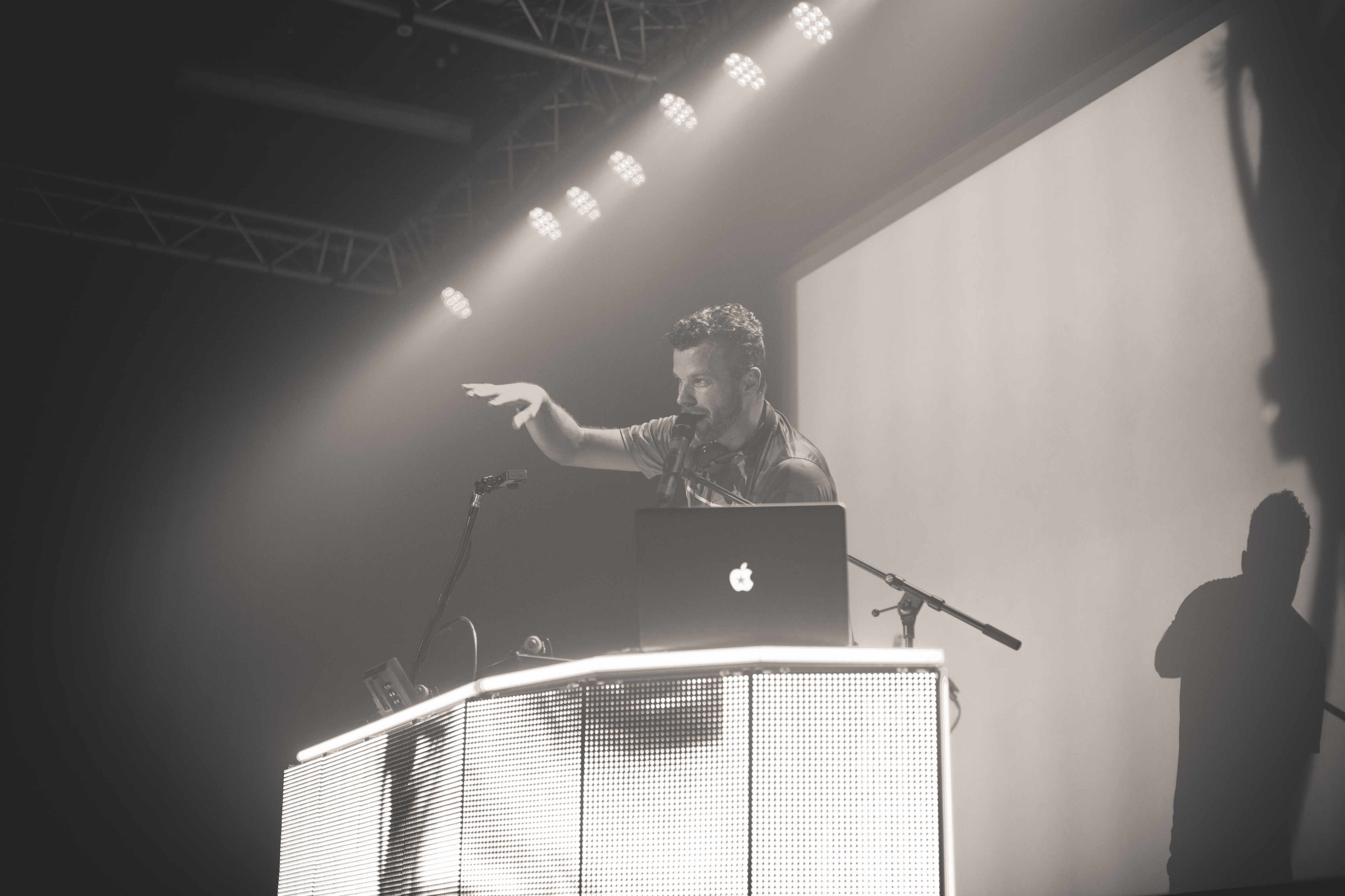 A DJ stands in a booth made of LED screens, gesturing at the crowd and talking into a mic.